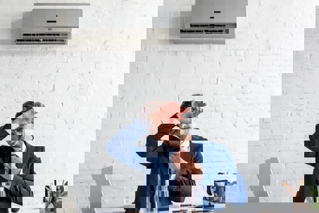 A hot, frustrated man in a suit wipes his forehead under an AC unit.