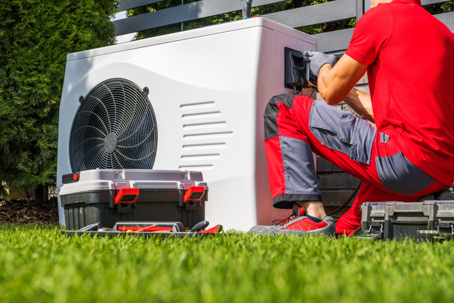 A technician in a red shirt services an outdoor heat pump unit on a green lawn.