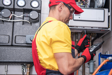 A technician in a red cap and yellow shirt inspects a furnace system, holding a diagnostic tool.