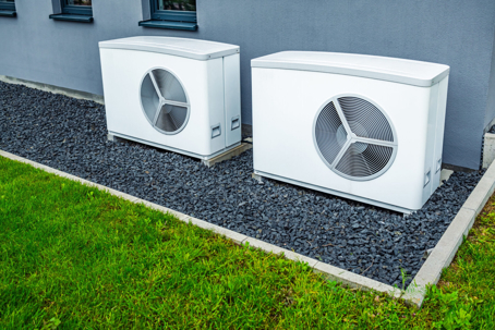 Two white outdoor heat pump units sit on dark gravel next to a gray house.
