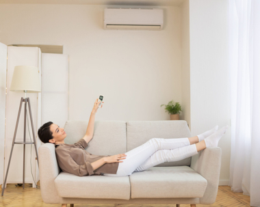 A woman relaxes on a light sofa, smiling as she uses a remote to adjust the wall-mounted air conditioner above her.