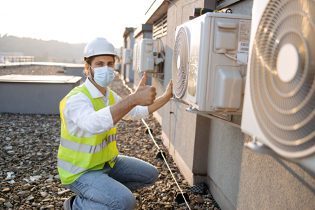A masked technician in a hard hat and safety vest kneels on a rooftop next to outdoor AC units, giving a thumbs-up.