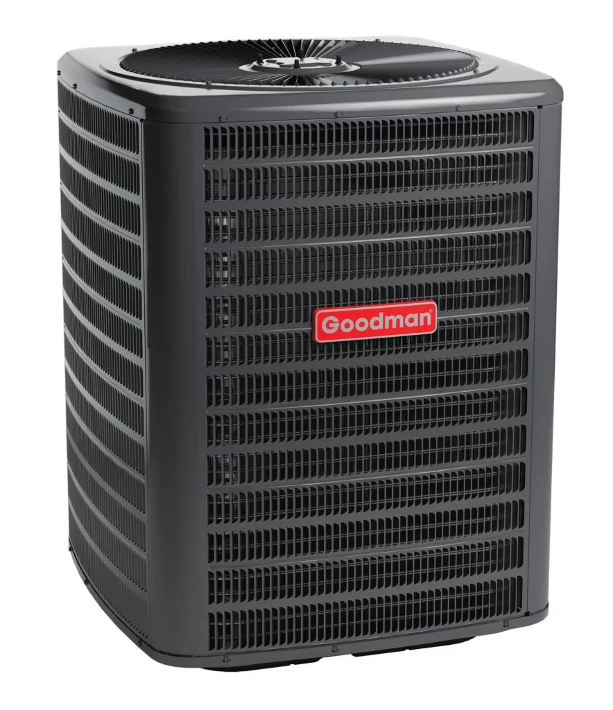A dark gray Goodman outdoor AC unit with a fan on top and grates on its sides. A red oval label displays "Goodman" in white text.